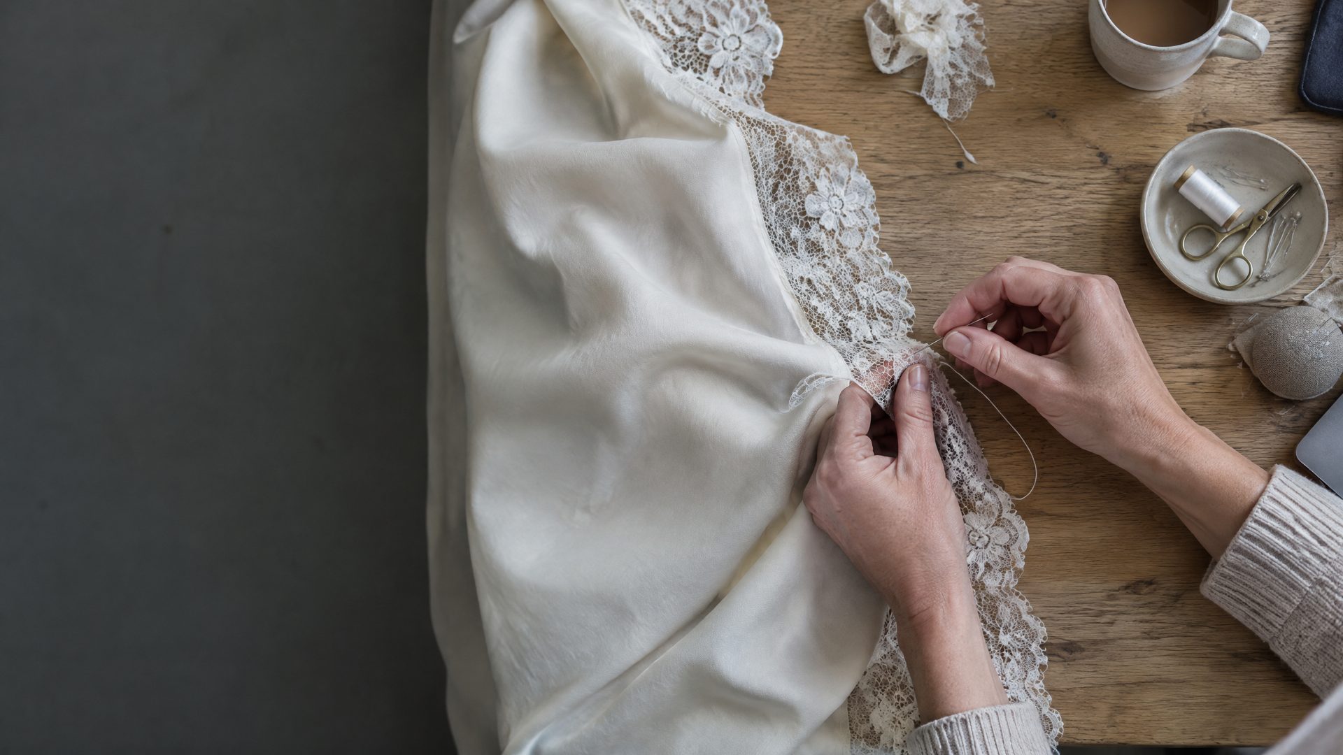 Overhead close-up of hands stitching the lace edge of ivory silk on a wooden table, with a dish of thread and scissors and a cup nearby.