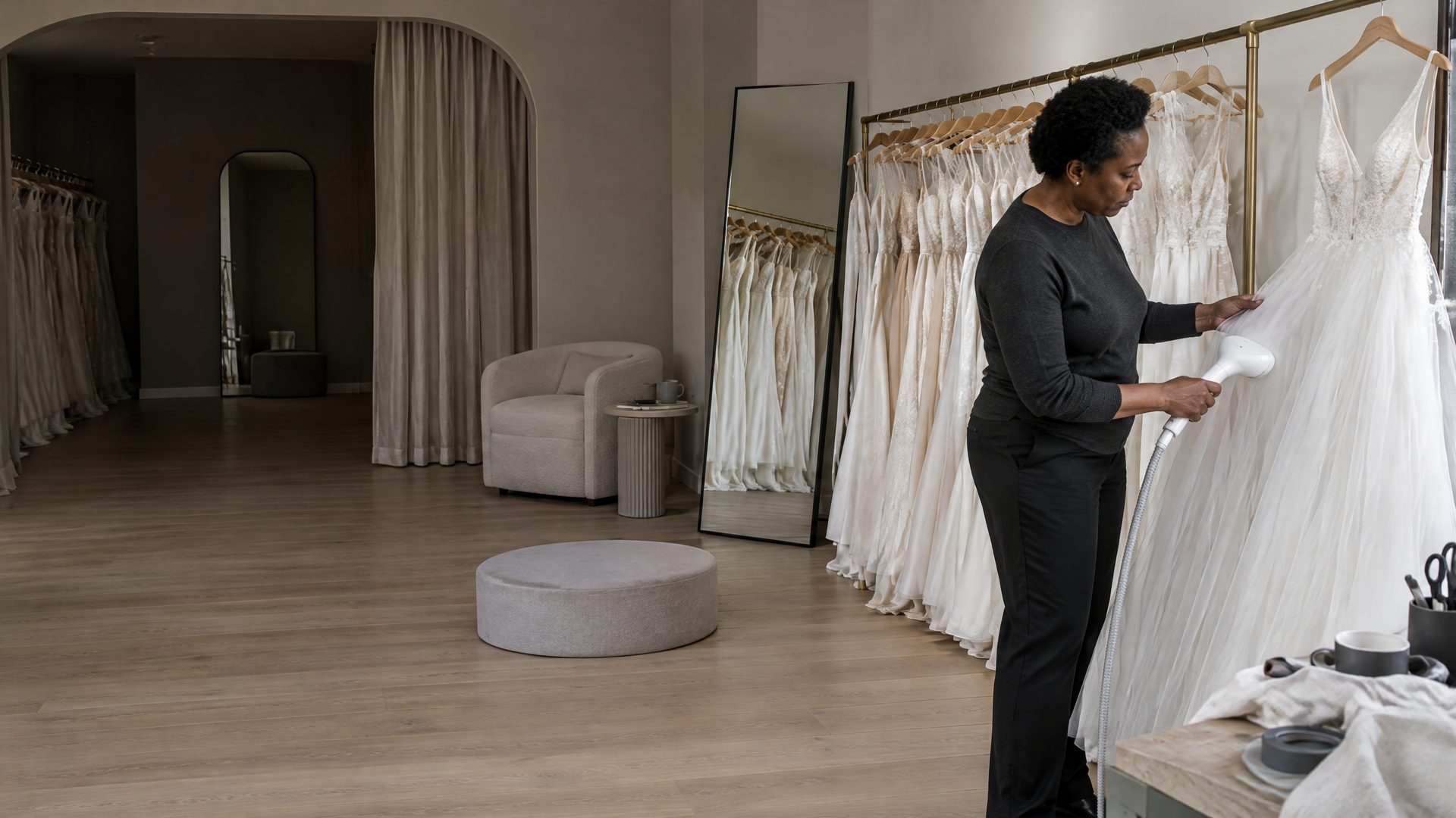 In a bridal boutique, a woman in a charcoal jumper steams the tulle skirt of a wedding gown on a brass rail, with a fitting podium, armchair and full-length mirror behind her.